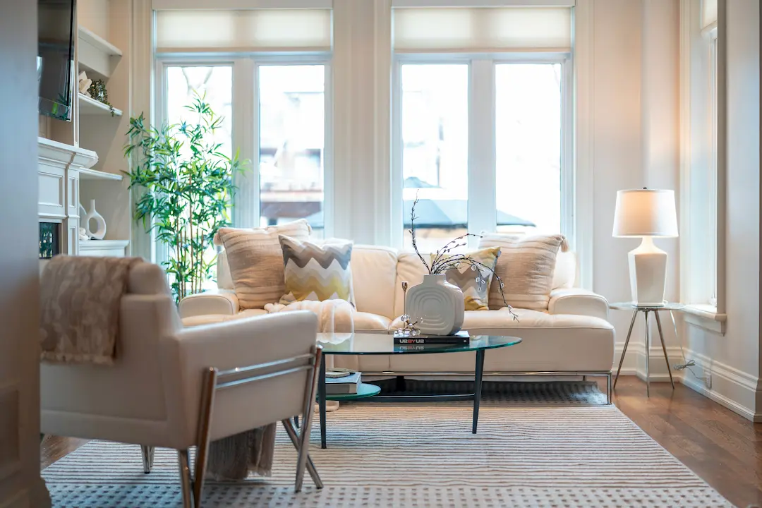 Jasny, elegancki salon z kremową sofą, fotelami, szklanym stolikiem kawowym, wysokimi oknami, roślinami i lampą podłogową; panuje tu ciepła, przytulna atmosfera.